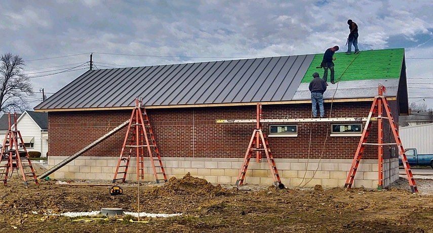 Construction workers installing a metal roof on a small brick building using red ladders and support beams.